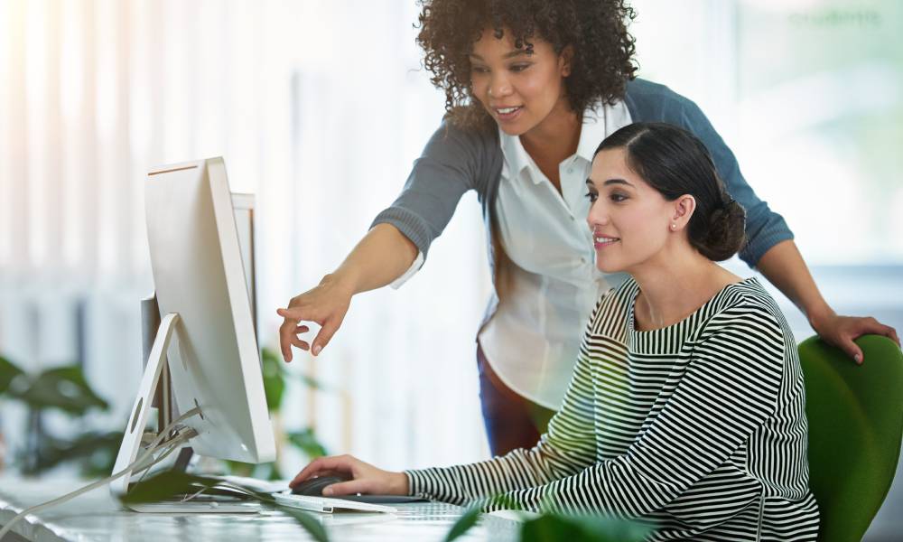 A female executive and her employee smiling as the executive points to the employee's computer to explain something.