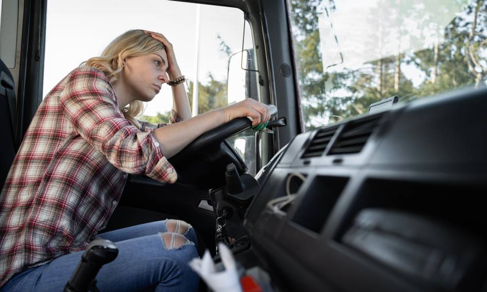 A woman in a plaid shirt sits in a truck with one hand on the wheel and one on her head. She is frowning.