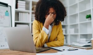 A black woman in a yellow blazer and striped dress shirt pinches the bridge of her nose, appearing in pain while at a desk.