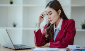 An Asian woman brushing her finger against her brow as she stares down at her work laptop, looking stressed.