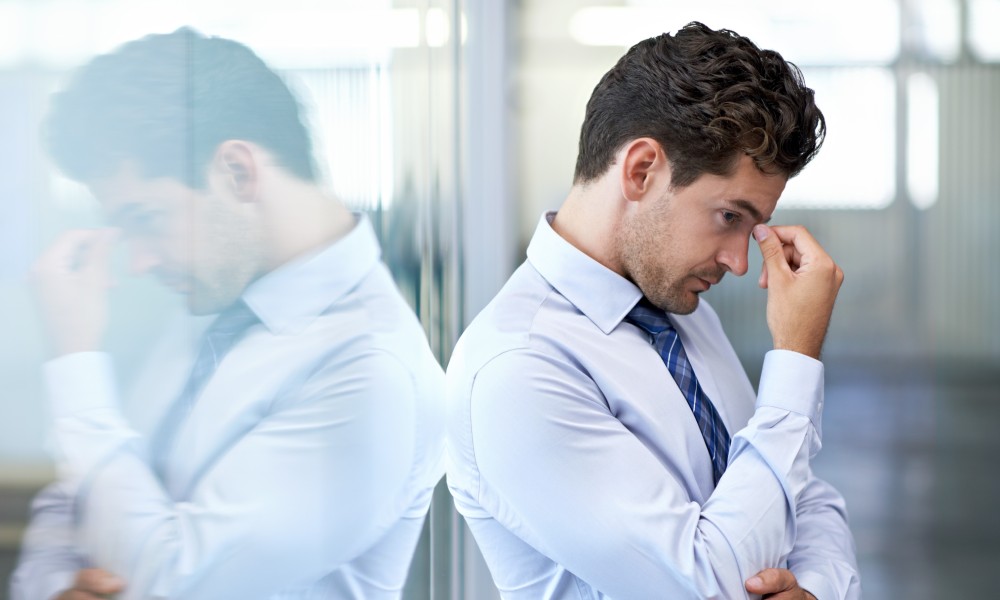 A well-dressed man in a blue dress shirt leaning against an office window and pinching his brow from stress.