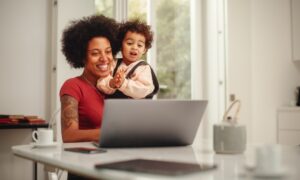 A woman sitting at a dining room table and holding her young child while they both look at her work laptop together.
