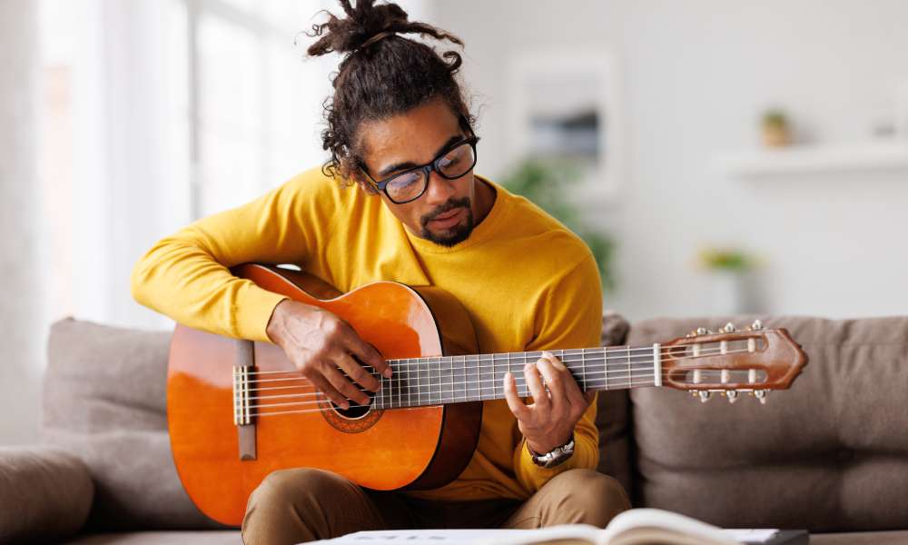 A man in a yellow shirt and black glasses is playing an acoustic guitar while sitting on the couch.