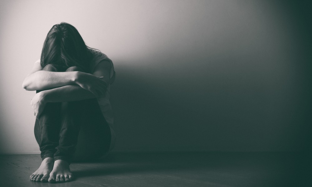 A young woman with long, dark hair sits on the floor with her arms wrapped around her legs. Her head rests on her knees.