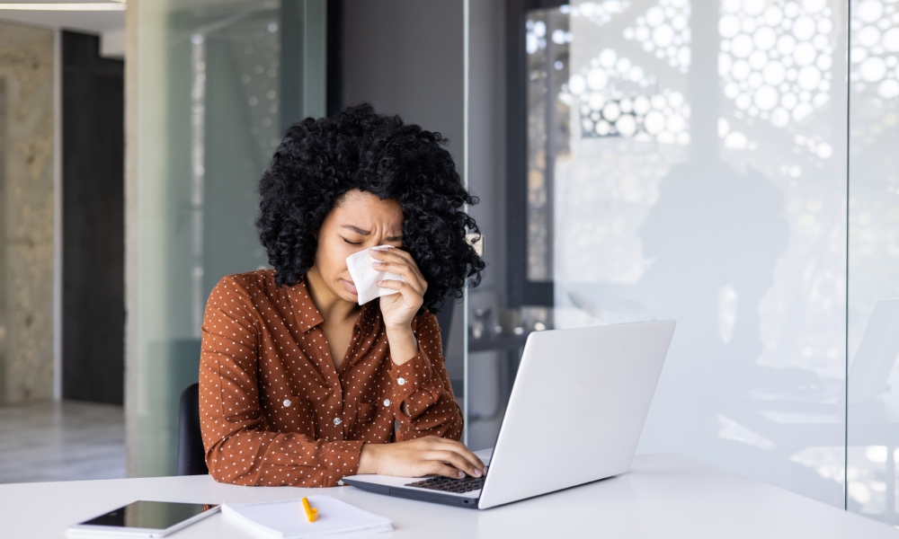 A young woman in a polka-dot dress shirt crying with a tissue at her desk in front of her computer in an office.