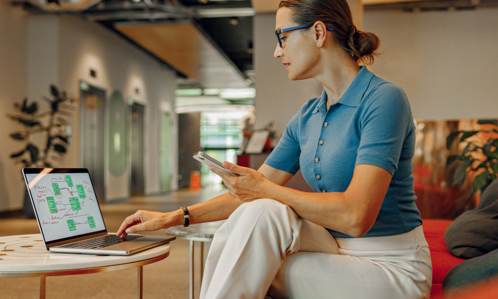 A young professional sitting in an open office setting and using a community couch as they work on their laptop.