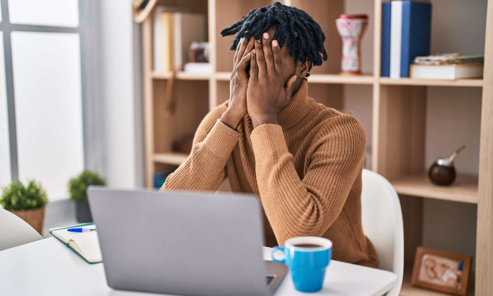 A young person in a light brown sweater sitting at their office desk and holding their face in their hands.