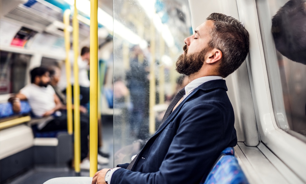 A tired man in business professional attire resting his head against the window of the train he’s riding on.