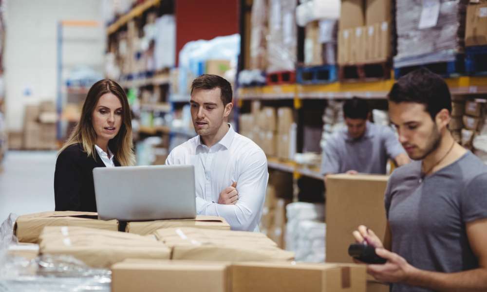 Two professionals looking over information on a laptop while standing in a warehouse aisle as others work around them.