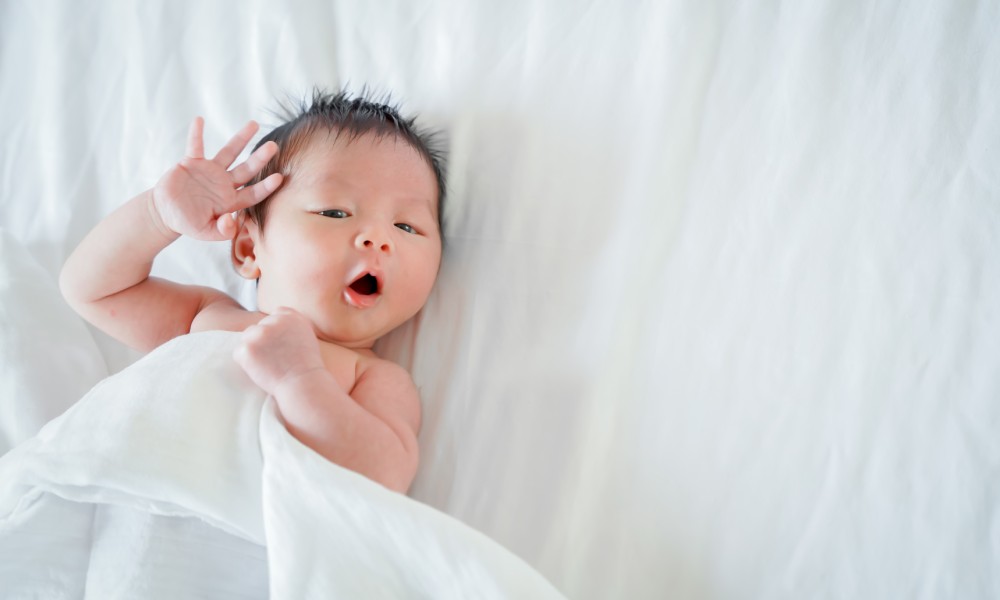 A newborn baby with dark hair is lying on a white sheet and reaching. His other hand is gripping the top white sheet.