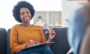A young woman softly smiling to someone off camera as she sits on a couch and writes information on a clipboard.