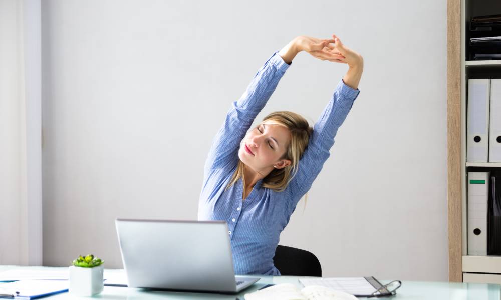 A woman sitting at a desk with a laptop, potted plant, and notebook. She stretches her arms over her head.