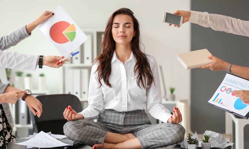 A woman sitting in a zen-like position on top of a table. There are hands reaching out to her with work-related items