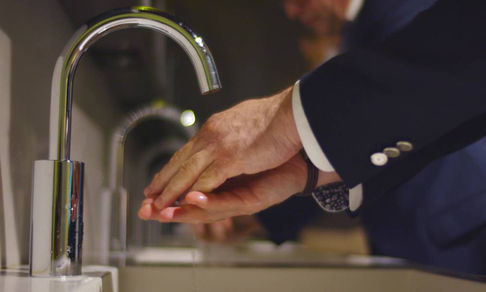 Two men wearing suits and washing their hands in a commercial restroom with stainless steel automatic touchless faucets.