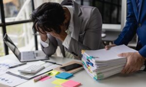A business professional holding their head in their hands while sitting at their desk as someone brings them more work to do.