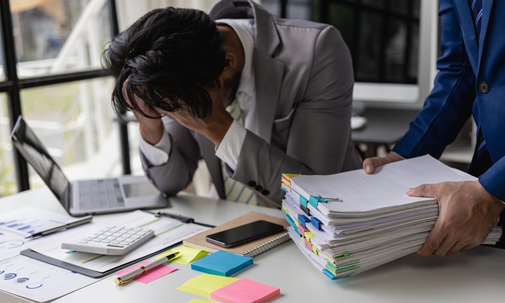 A business professional holding their head in their hands while sitting at their desk as someone brings them more work to do.