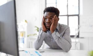 A young professional resting their hand in their hands while looking exhausted and staring at a large work monitor.