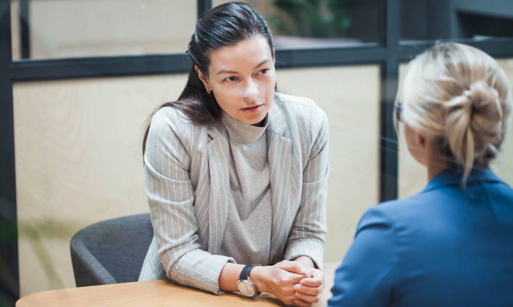 A business professional with a serious expression speaks to another professional across the table in a closed office.