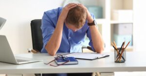 A man sitting at his work desk in front of a clipboard with his face down and his hands over his head.