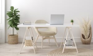 A white desk sits in the middle of a room with books, a silver laptop, and a tiny plant on top. A creme chair is at the desk.