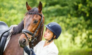 A young woman wearing a black riding helmet and white shirt stares at a brown horse. She's holding its face with her hand.