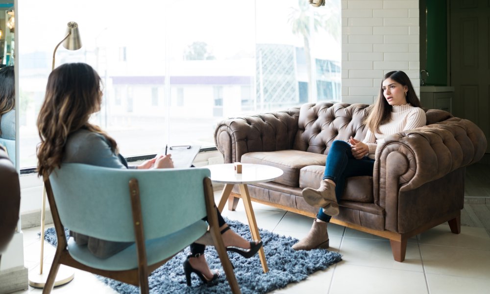 A woman sits on a brown leather couch across from a woman sitting on a wooden chair with blue upholstery.