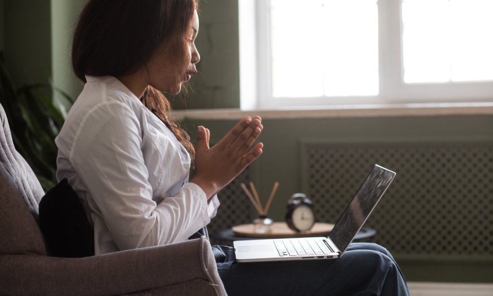 A therapist sitting in her chair with her laptop in her lap. She's talking with her hands with a patient on the call.