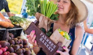 A smiling woman walking by a produce stand, holding a wooden box full of fresh peppers and kale that reads "Farmers Market."