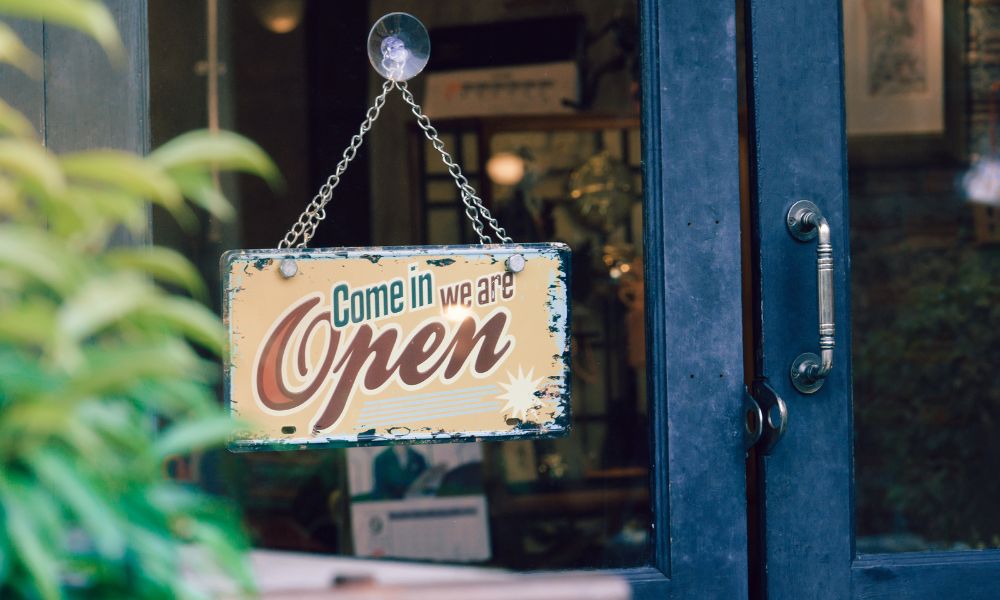 A small sign hanging by a chain inside a blue business door with a window reads "come in we are open".