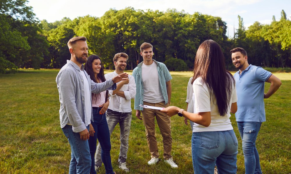 A group of people wearing casual clothing smiles as they all stand outside on the grass in a circle formation.