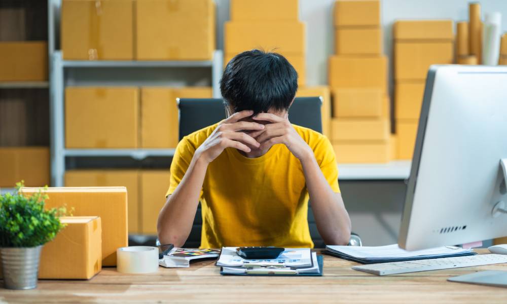 A young business owner sitting at a wooden desk and holding their head in their hands. Shelves with boxes are behind them.