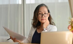 An older woman holding a set of papers in one hand and focusing on the screen of her open laptop while at a desk.