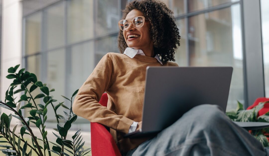 A woman wearing glasses smiles as she sits outside in a chair by a plant, with her laptop in front of her.