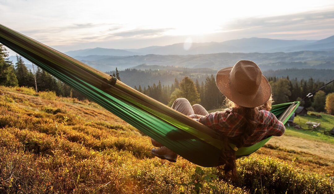 A person with a straw hat is relaxing on a green hammock overlooking a hill with trees and mountains in the distance.