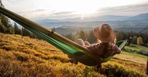 A person with a straw hat is relaxing on a green hammock overlooking a hill with trees and mountains in the distance.