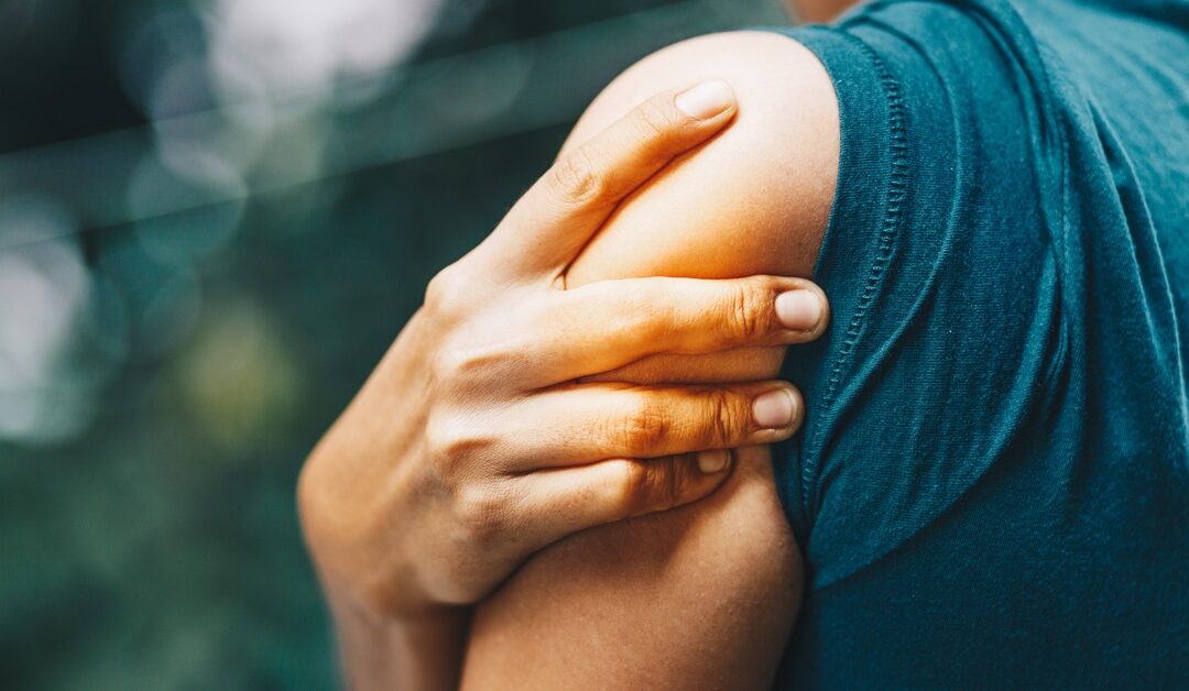 A person in a blue tank top clutching their upper left arm with their right hand due to pain while outdoors.