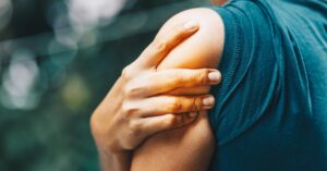 A person in a blue tank top clutching their upper left arm with their right hand due to pain while outdoors.