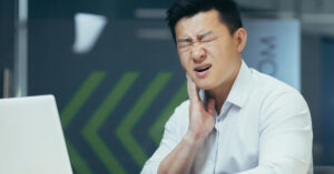 A man in a button-up shirt sitting in front of a laptop in his office. He is holding his jaw and making a pained expression.