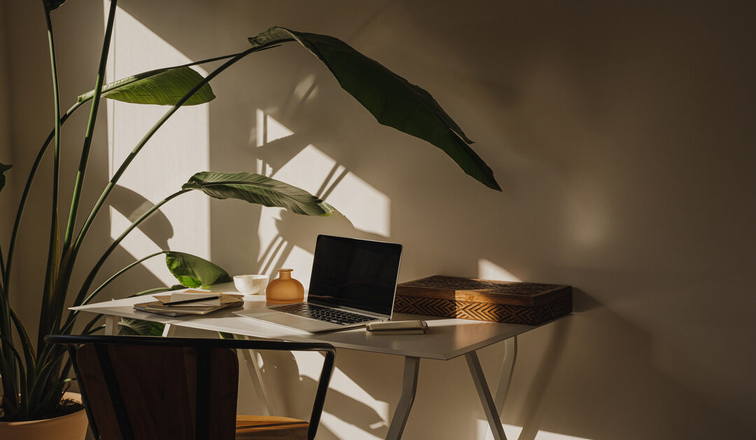 A small work desk with a laptop and notepads on the surface in a dark room illuminated by window light.