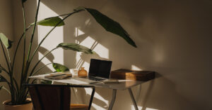 A small work desk with a laptop and notepads on the surface in a dark room illuminated by window light.