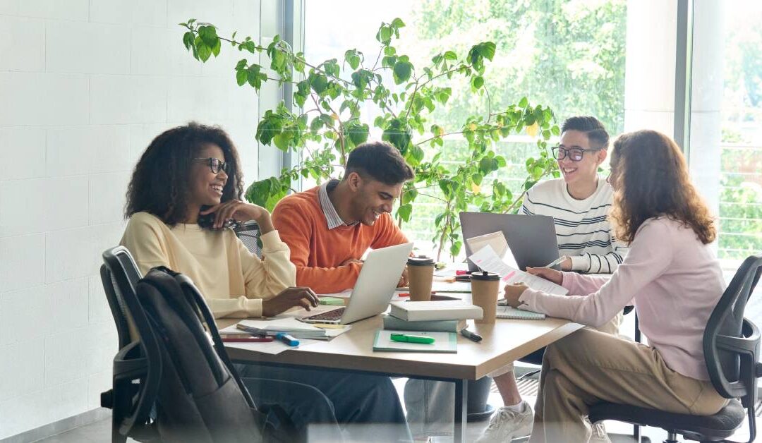 Four diverse coworkers sit around a table in a meeting room, working on laptops and overviewing papers while smiling.