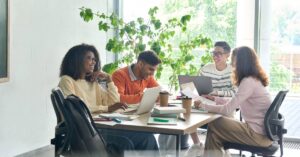Four diverse coworkers sit around a table in a meeting room, working on laptops and overviewing papers while smiling.