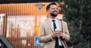 A smiling man in a suit stands outside, holding a drink and carrying a brown satchel. There's a building in the background.
