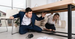 Two office workers crouching under a wooden table. The man leans on a chair, surrounded by scattered books and pencils.