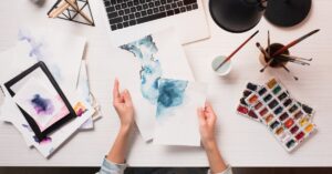 A person sitting at a desk with a laptop open but turned off. They hold papers with blue watercolor paint on them.