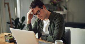 A stressed man leaning against his home desk and staring down at his open laptop with a tense expression.
