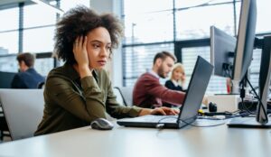 A woman working in an office puts her hand on her head while looking at her laptop and computer monitor.