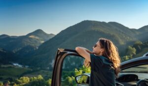 A person with long hair leaning against their open car door. Tall, tree-covered mountains surround them.