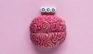 A plastic brain with swirls on its surface sitting on a pink background beside blocks that read "OCD".