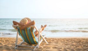 A woman wearing a woven sunhat sits in a teal striped chair on the beach, facing the ocean and the sun.